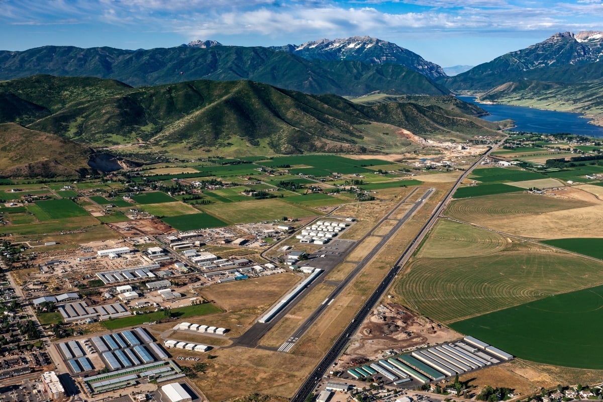 Aerial view of a rural airport runway with surrounding farmland, industrial buildings, and mountains in the distance
