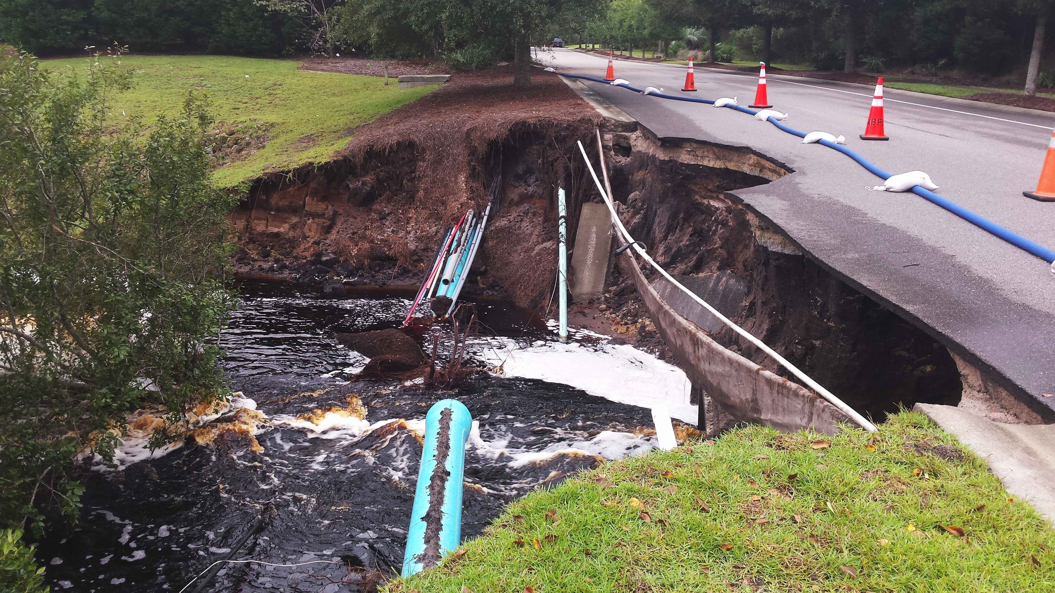 Washed-out roadway with severe erosion exposing pipes beneath the pavement, water flowing below, and traffic cones marking the damaged road edge