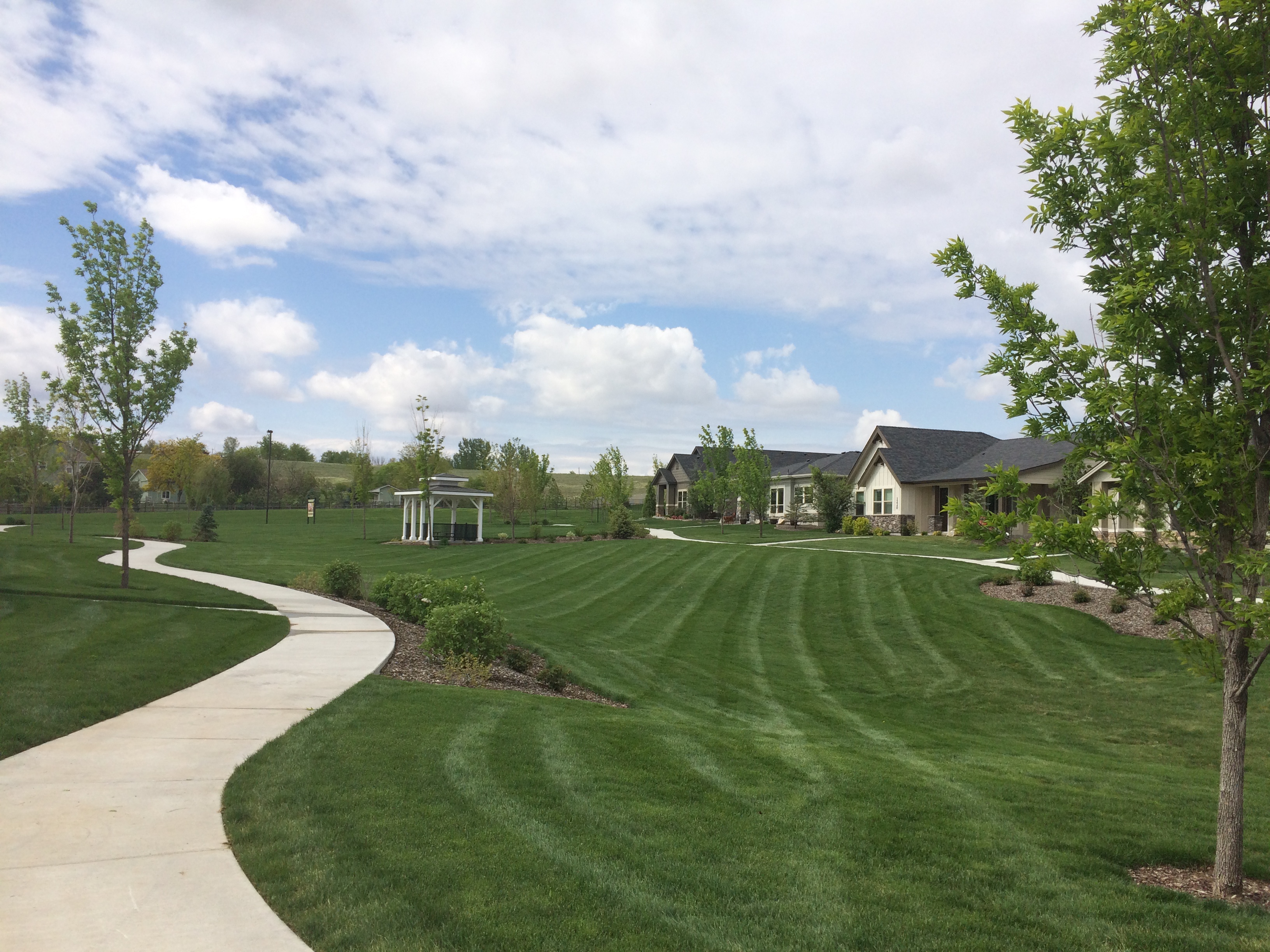 Curved walkway through a landscaped park with green lawn, trees, gazebo, and nearby houses
