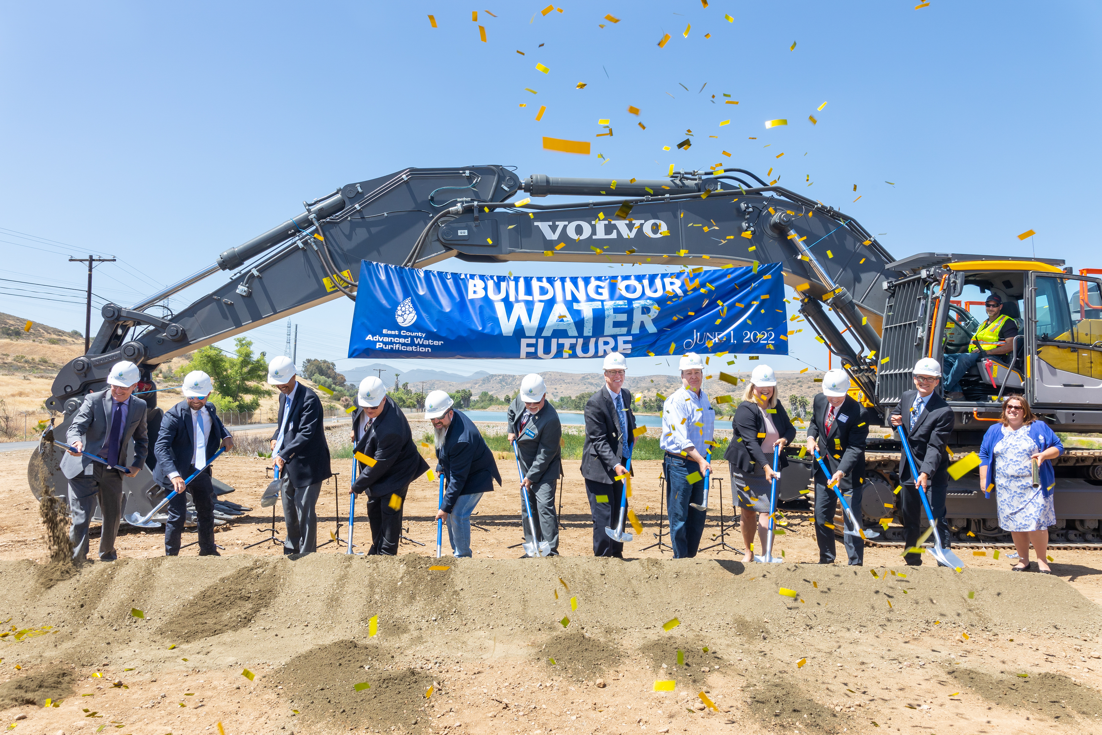 Groundbreaking ceremony with officials using shovels in front of an excavator and banner reading Building Our Water Future with confetti falling