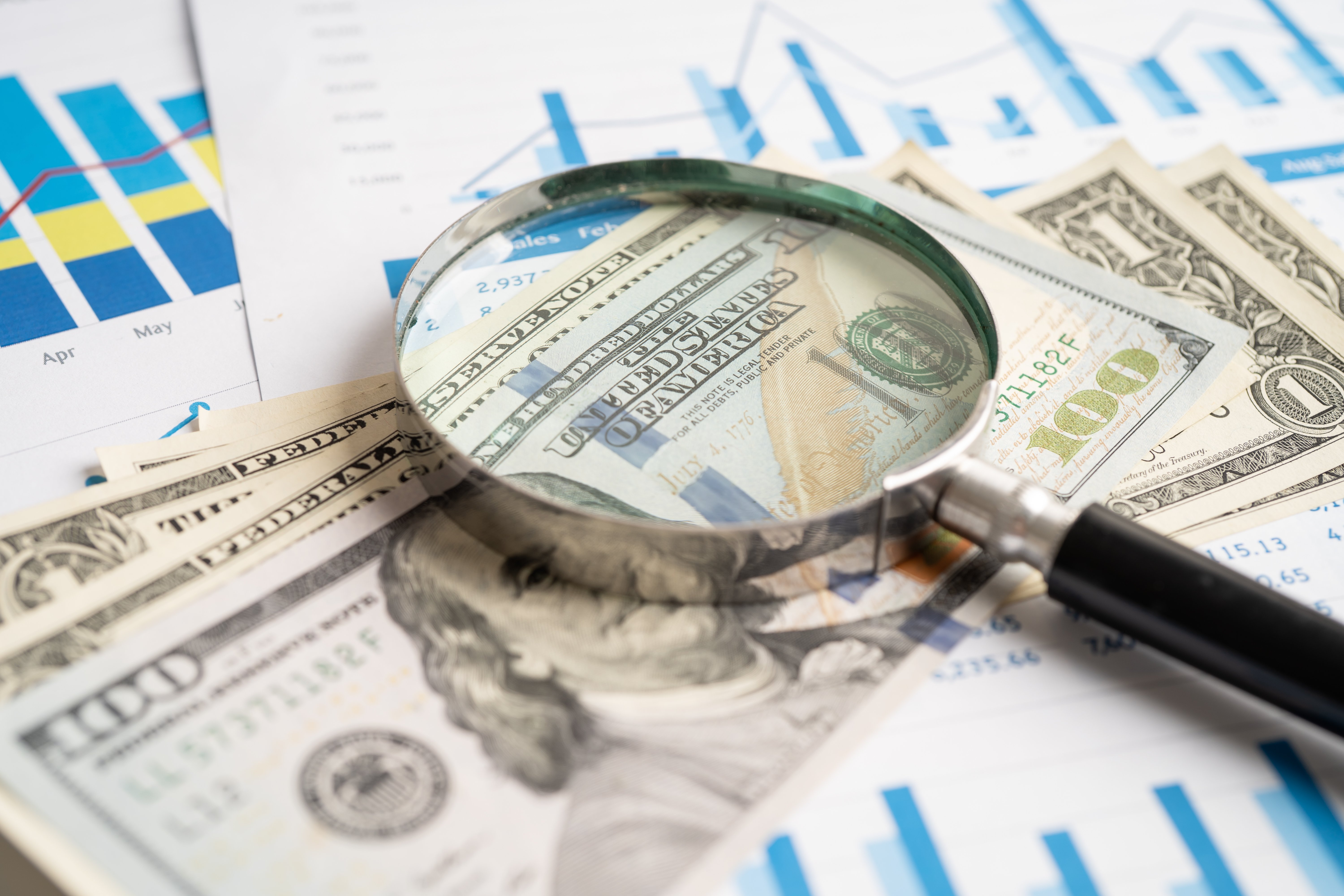 Magnifying glass over US dollar bills and financial charts showing graphs and data